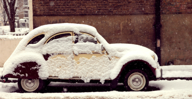 Une voiture de collection recouverte de neige stationnée sur le bord de la chaussée devant un mur d’immeuble en briques rouges.