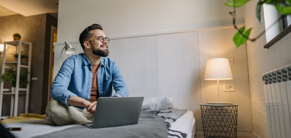 Jeune homme dans son appartement, assis confortablement sur son lit dans une chambre confortable et bien éclairée, recherchant une assurance locataire sur son ordinateur portable.
