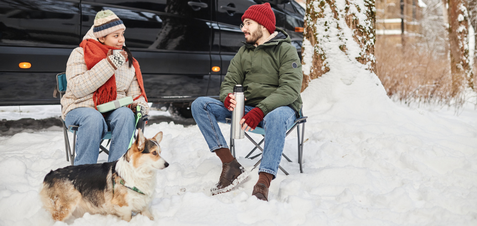 Un couple assis sur des chaises dans un camping d’hiver avec leur chien devant leur VR.  