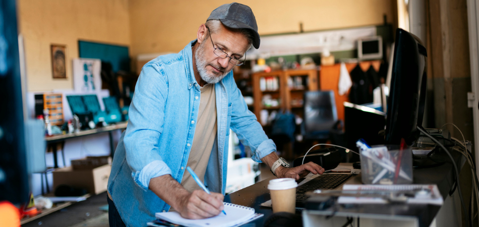 Un homme d’âge mûr est dans son atelier, en train de prendre des notes et de planifier les tendances en matière d’assurance des entreprises pour 2026.  