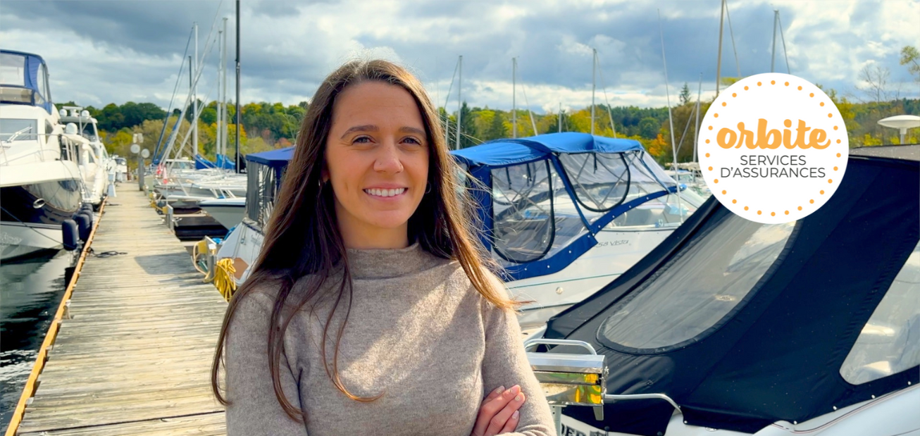 Une personne aux cheveux longs se tient debout sur un quai et parle d’assurance marine. Elle sourit et porte un chandail beige tandis que plusieurs bateaux sont amarrés derrière elle.