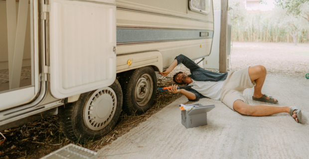 Un homme portant des vêtements d’été est allongé par terre avec un outil à la main; il est en train de réparer le pneu de sa roulotte. 