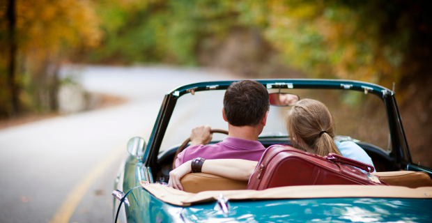 Un couple se promène en voiture par une belle journée chaude. La voiture de collection décapotable turquoise à l’intérieur beige est impeccable. Une valise bourgogne est visible sur la banquette arrière.