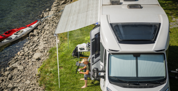 Une autocaravane moderne de catégorie C garée au bord d’un lac avec l’auvent sorti; le conducteur est assis, en train de prendre un café.  