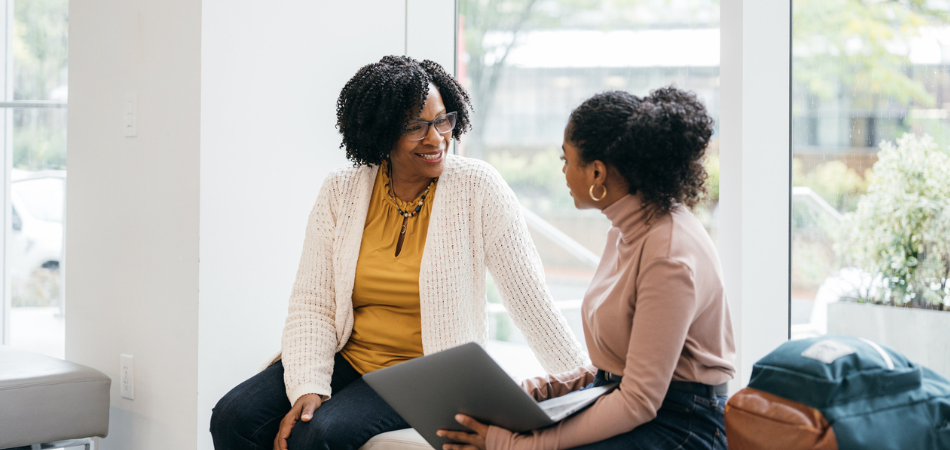 Deux personnes assises sur un banc dans un espace intérieur lumineux et moderne avec de grandes fenêtres. L’une d’elles tient un ordinateur portable et elles semblent discuter de l’assurance locataire.  