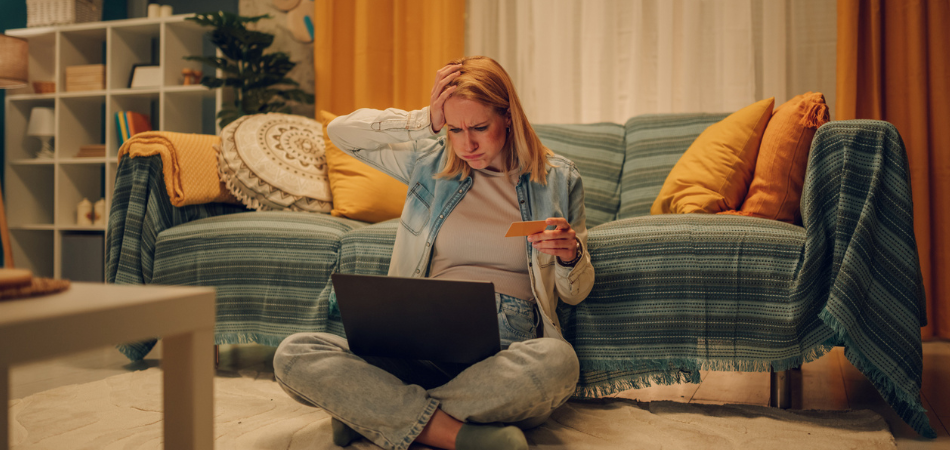 Une personne est assise sur le sol de son salon, tenant une carte de crédit et regardant son ordinateur portable avec inquiétude, ce qui suggère qu’elle a peut-être été victime d’une arnaque liée aux achats en ligne.