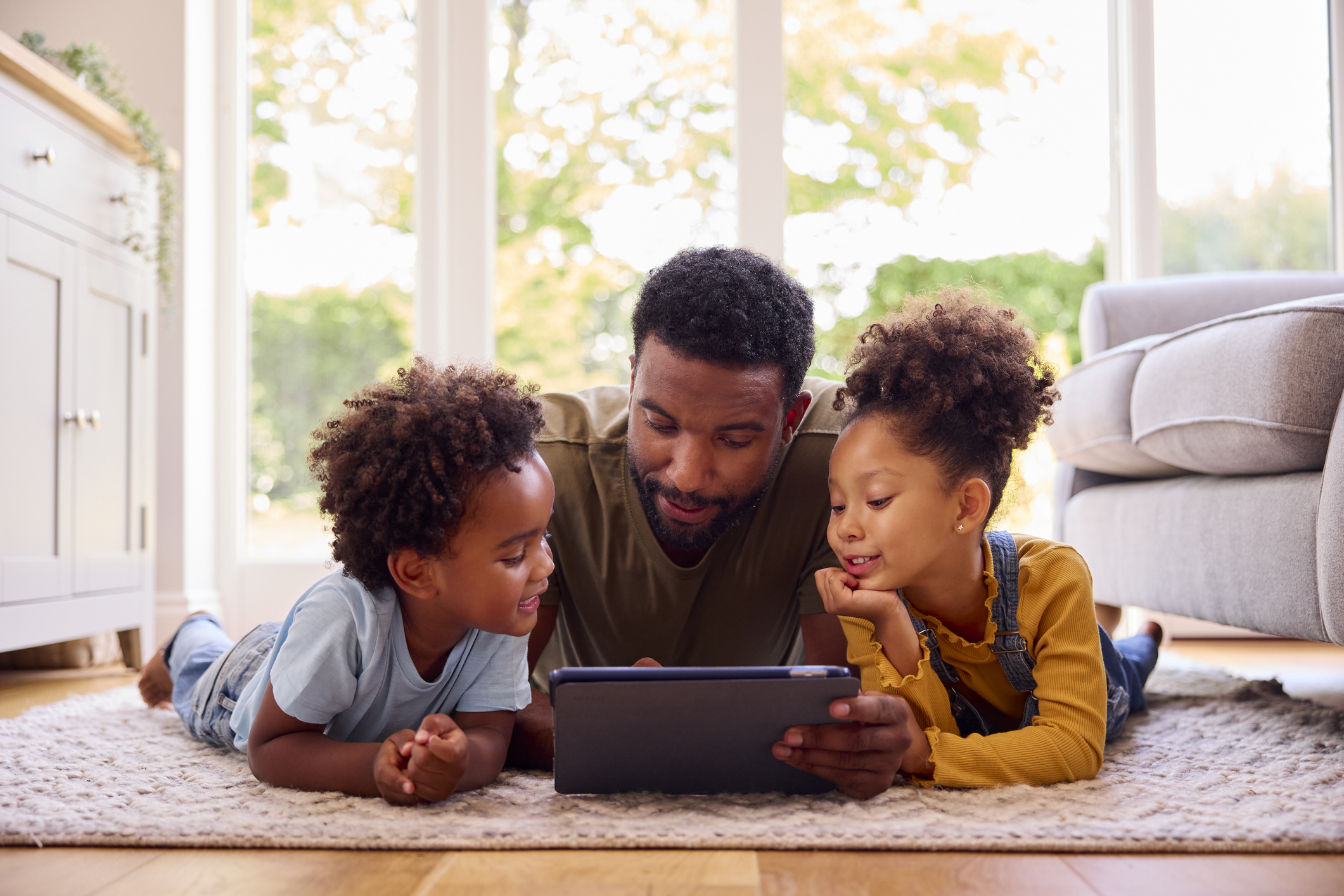 Un père et ses deux enfants se prélassent sur un tapis en utilisant une tablette