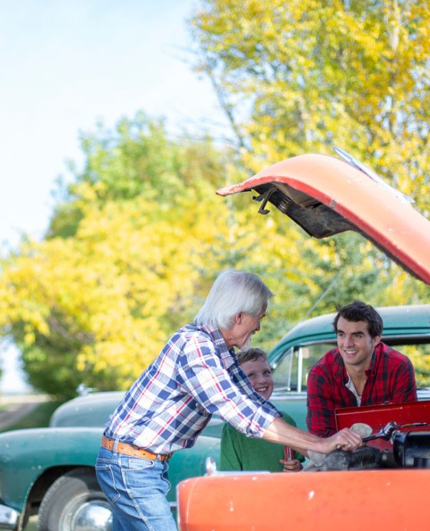 Un grand-parent travaille sous le capot d'une voiture classique pendant que son fils et son petit-fils la regardent.