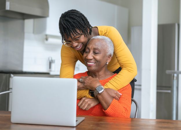 Une adolescente noire embrasse sa grand-mère, qui travaille sur un ordinateur portable à la table de la cuisine, par derrière.