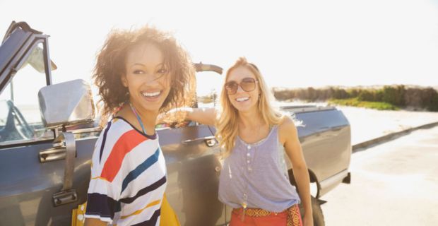 Two women, taking a break from their road trip, are standing beside their car on a sunny day, laughing and smiling.