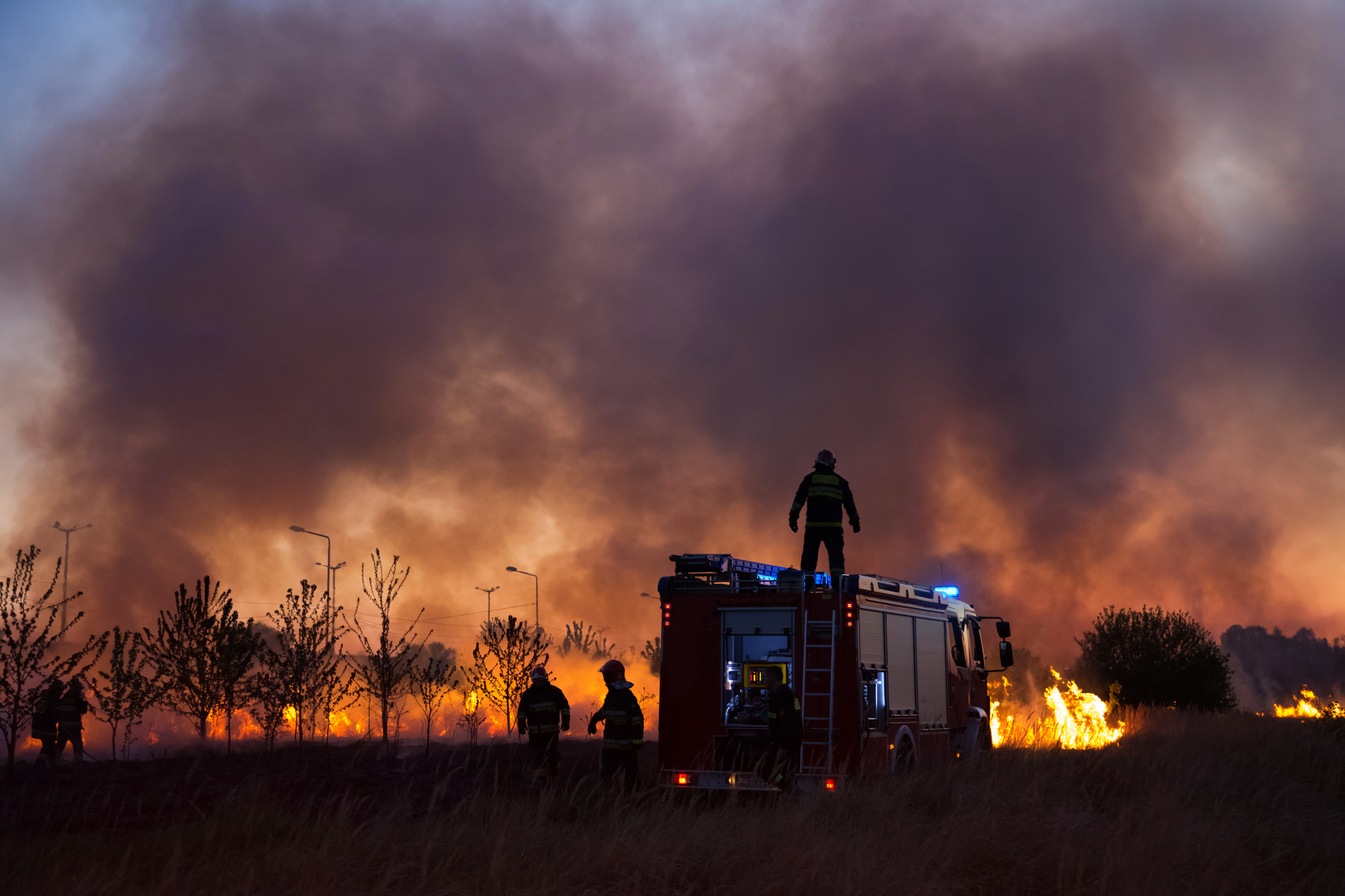Pompiers debout autour d’un camion de pompiers avec des incendies de forêt et un ciel enfumé en arrière-plan. 