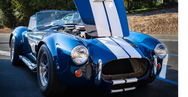 Une splendide Ford Shelby Cobra 427 de 1967 est garée sur une route. La voiture, dont le capot est ouvert, est d’un bleu métallique étincelant avec des bandes blanches dessus. Le capot s’ouvre à partir de la base du pare-brise. C’est véritablement une magnifique voiture de collection.