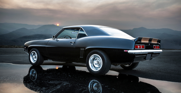 Une Chevrolet Camaro noire est garée sur une route de gravier près des montagnes.