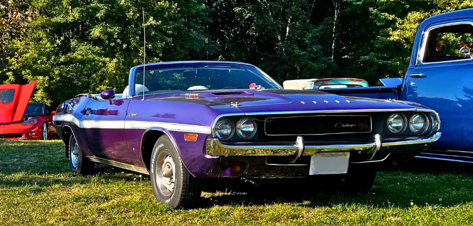 Une Dodge Challenger R/T décapotable violette classique garée sur l’herbe lors d’un salon automobile en plein air, photographiée depuis l’angle avant gauche.