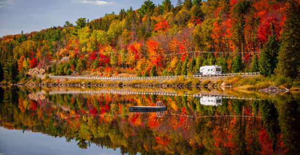 Une caravane roule près d'une forêt aux couleurs automnales. On aperçoit le reflet de l'autoroute 60 sur le lac de Deux Rivières. Parc Algonquin, Ontario, Canada.