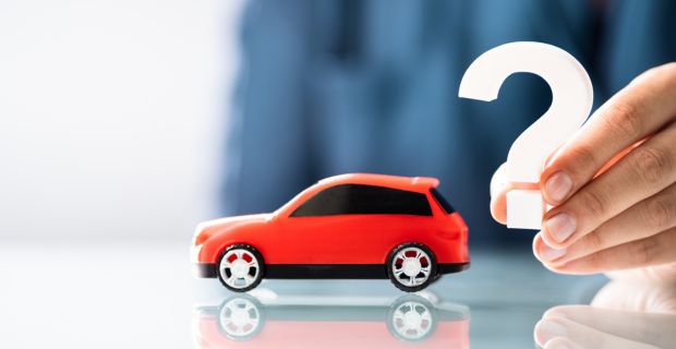 Une petite voiture rouge posée sur une table avec une personne tenant un point d’interrogation derrière la petite voiture.