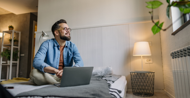 Jeune homme dans son appartement, assis confortablement sur son lit dans une chambre confortable et bien éclairée, recherchant une assurance locataire sur son ordinateur portable.