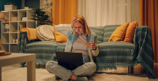 Une personne est assise sur le sol de son salon, tenant une carte de crédit et regardant son ordinateur portable avec inquiétude, ce qui suggère qu’elle a peut-être été victime d’une arnaque liée aux achats en ligne.